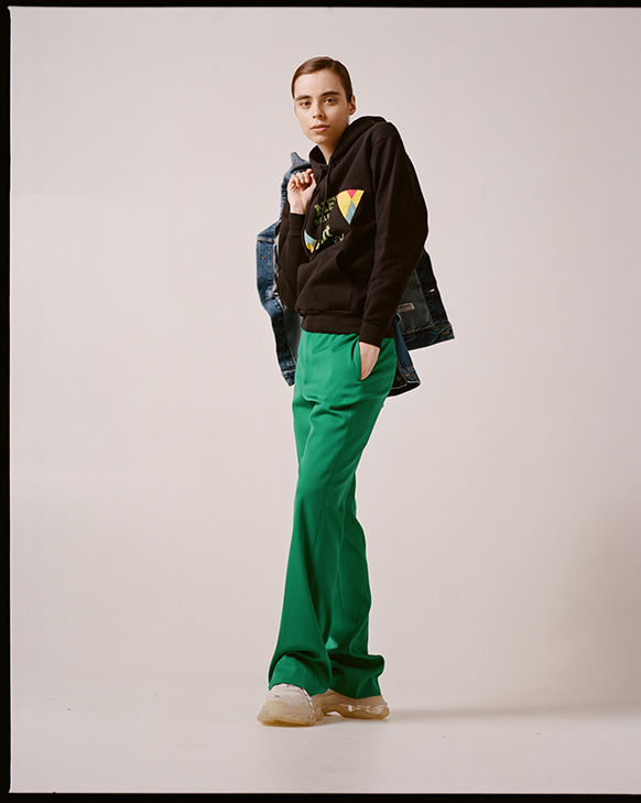 Female model wearing Balenciaga denim jean jacket with upcycled quilted patches, black sweatshirt with triangle applique, green pants and white Triple S sneakers. 