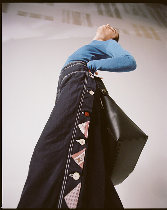 Female model wearing Stella McCartney women's wide-leg pants with patchwork upcycled details, blue sweater, and black vegan leather bag.