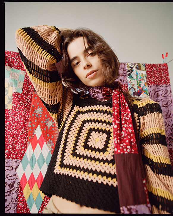 Female model wearing Ulla Johnson knitwear patterned sweater with scarf made from upcycled dresses. Tapestry quilt hanging behind her also made of upcycled dress fabrics. 