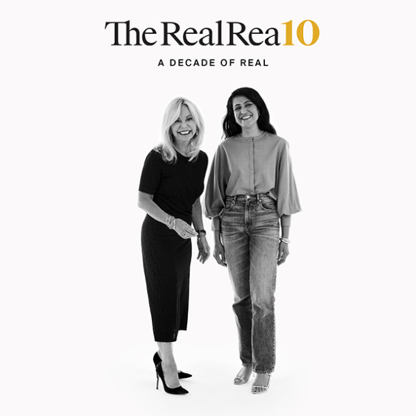 The RealReal founders Julie Wainwright, founder and CEO, and Rati Sahi Levesque, president. Standing together on a white background laughing and smiling. Julie is wearing a black dress and black pumps. Rati wears a grey top with bell sleeves, pale denim jeans, and white strappy sandals.