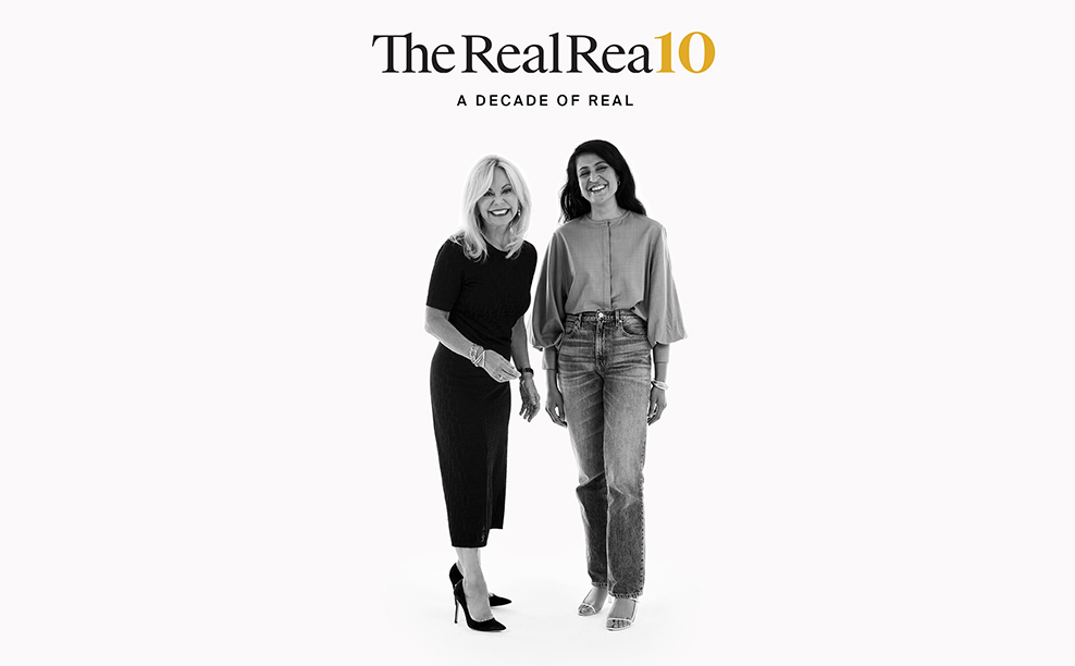 The RealReal founders Julie Wainwright, founder and CEO, and Rati Sahi Levesque, president. Standing together on a white background laughing and smiling. Julie is wearing a black dress and black pumps. Rati wears a grey top with bell sleeves, pale denim jeans, and white strappy sandals.