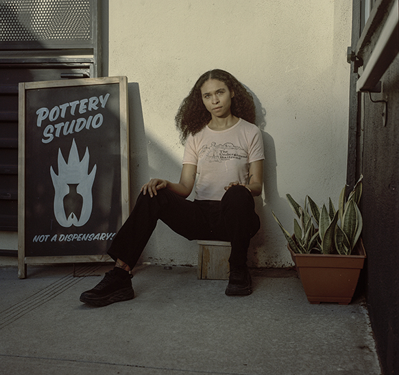 Artist Anjelica Alubowicz sitting on the sidewalk next to a plant and a sign that says "Pottery Studio, Not a Dispensary"