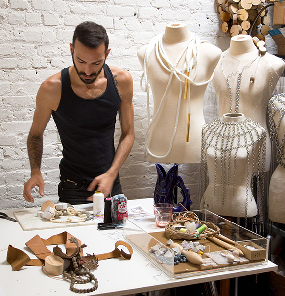 Jewelry designer Edgar Mosa in his studio with goldsmithing tools on the table, and three mannequins next to him wearing chains, jewelry and accessories. 