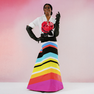 Miss J Alexander stands in a rainbow striped full-length skirt by Christopher John Rogers, a white button down shirt with a red rose printed on the front, and long, black gloves in front of a pink ombre background.