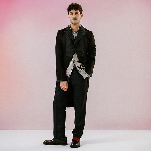 For pride prom, Alex Tudela stands in front of a pink ombre background, wearing a black suit jacket and trousers with a silver, lame button down shirt, untucked. 