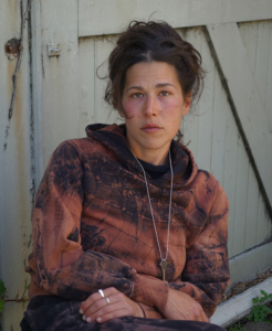 Artist Amy Giovanna Rinaldi sitting in front of a barn door wearing one of her pribnted "queer camo" sweatshirts.