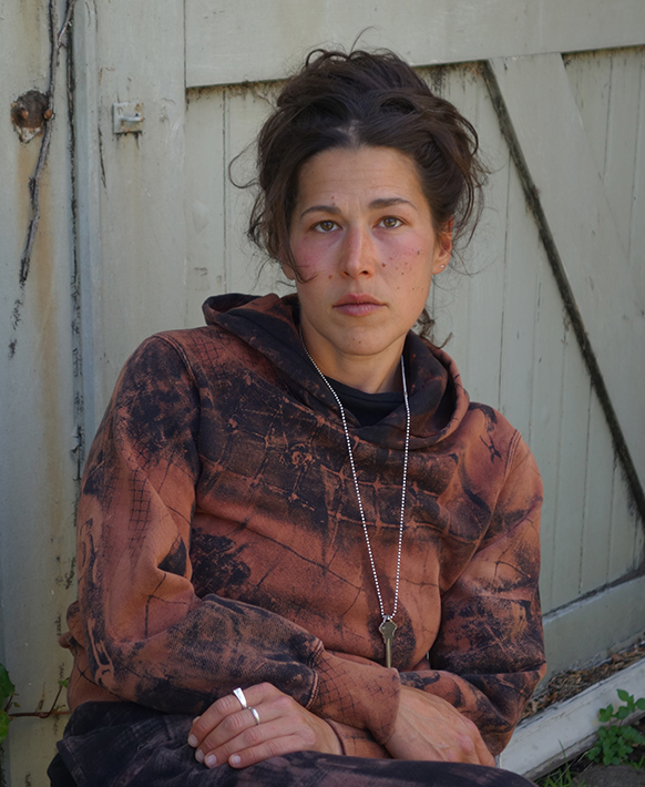 Artist Amy Giovanna Rinaldi sitting in front of a barn door wearing one of her pribnted "queer camo" sweatshirts. 