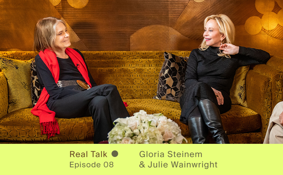 Gloria Steinem and Julie Wainwright sitting on a sofa talking, smiling and laughing together. Both wearing black clothing.