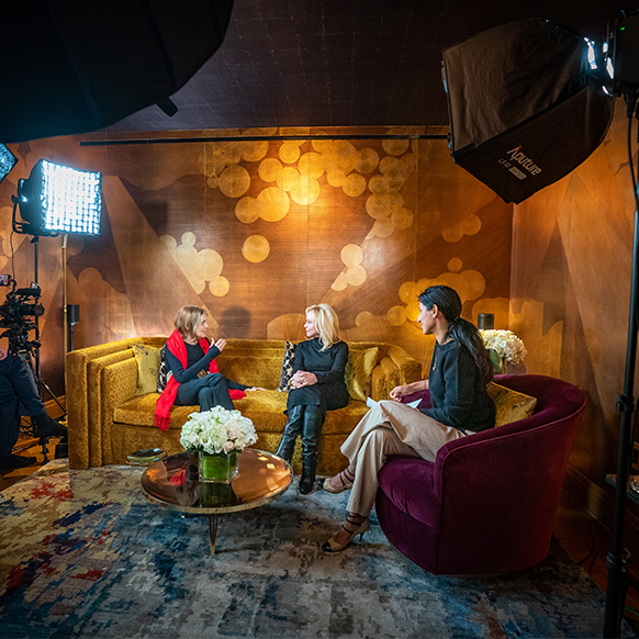 Gloria Steinem in a red scarf, black fashion clothing, and a belt and Julie Wainwright wearing a black dress, black leather boots, gold bracelets and jewelry. two women sitting on a couch looking at each other, talking and smiling. with host Rita Nakouzi on the right and camera equipment surrounding them. 
