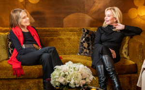 Gloria Steinem in a red scarf, black fashion clothing, and a belt and Julie Wainwright wearing a black dress, black leather boots, gold bracelets and jewelry. two women sitting on a couch looking at each other, talking and smiling.