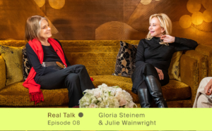 Gloria Steinem in a red scarf, black fashion clothing, and a belt and Julie Wainwright wearing a black dress, black leather boots, gold bracelets and jewelry. two women sitting on a couch looking at each other, talking and smiling.