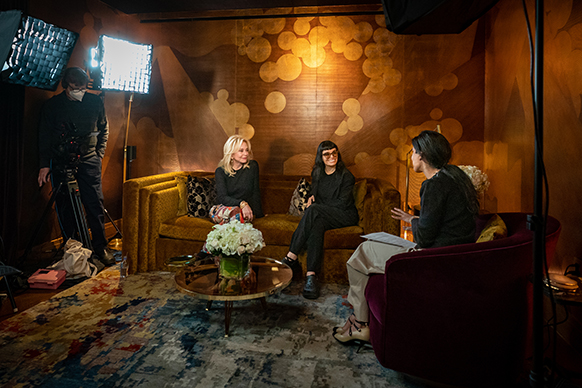 Julie Wainwright, Norma Kamali sitting on a sofa and Rita Nakouzi sitting in an armchair talking with camera equipment, lights, and crew members standing around them and a glass coffee table with flower arrangement in the center. Clothing: Julie wears a black top, Norma Kamali skirt, platform shoes, and jewelry. Norma Kamali wears a Norma Kamali black suit and black Crocs. Rita wears Chanel shoes, khaki pants and a black sweater. 
