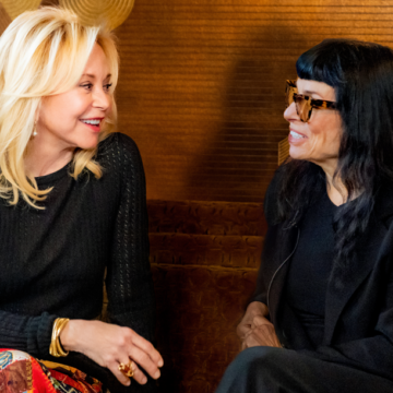 Julie Wainwright, Norma Kamali sitting on a sofa smiling, talking, and laughing. Clothing: Julie wears a black top, Norma Kamali skirt, platform shoes, and jewelry. Norma Kamali wears a Norma Kamali black suit, black framed eyeglasses and black Crocs.