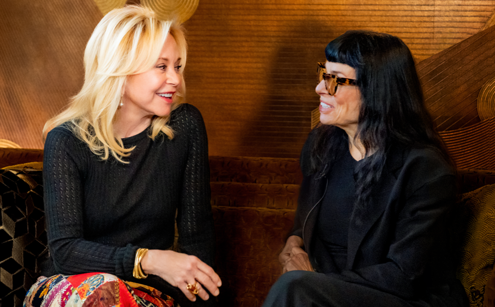 Julie Wainwright, Norma Kamali sitting on a sofa smiling, talking, and laughing. Clothing: Julie wears a black top, Norma Kamali skirt, platform shoes, and jewelry. Norma Kamali wears a Norma Kamali black suit, black framed eyeglasses and black Crocs.